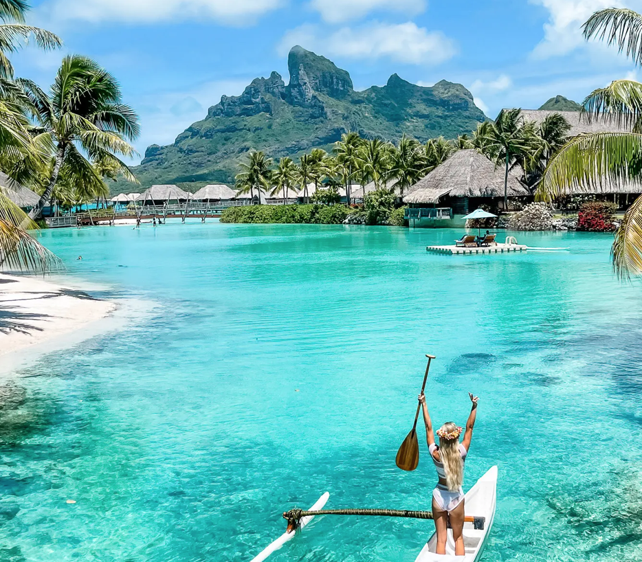 A person in a white swimsuit paddles in a canoe on clear turquoise water, surrounded by lush palm trees and mountains in the background.