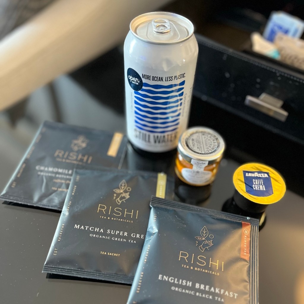 A selection of Rishi tea bags, a can of Still Water, a small jar of honey, and a Lavazza coffee capsule arranged on a table.