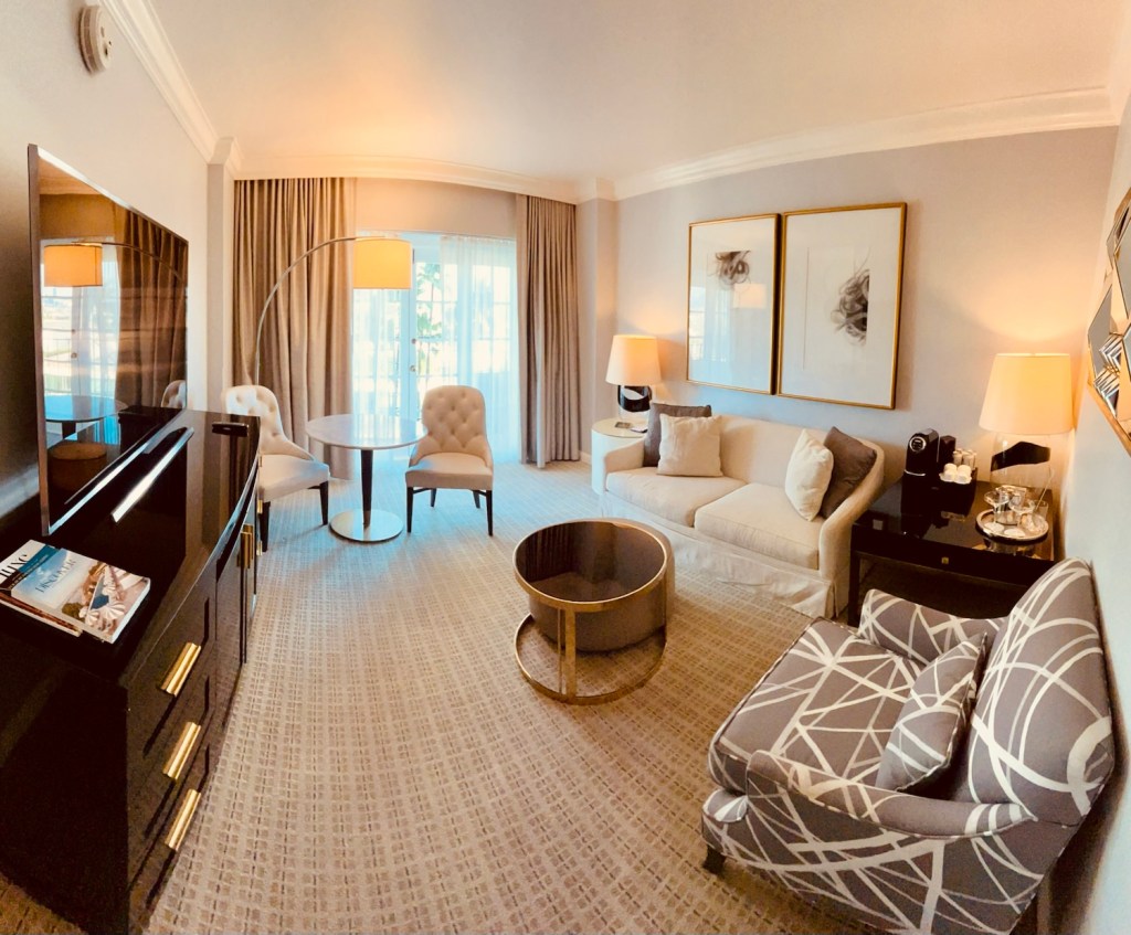 A luxurious hotel suite featuring a modern design with plush seating, a marble coffee table, and a large flat-screen TV. Large windows allow natural light to fill the room, showcasing elegant decor and soft furnishings.