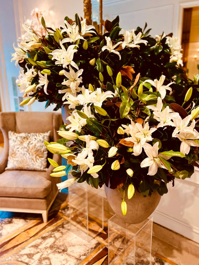 A large floral arrangement featuring white lilies and green foliage, displayed elegantly in a modern vase, next to a plush armchair in a luxurious hotel lobby.