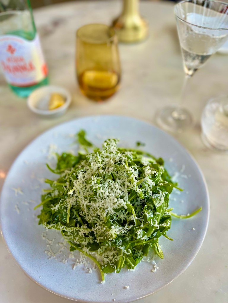 A top-down view of a plate filled with a fresh arugula salad topped with grated cheese, accompanied by a glass of water, a cocktail glass, and a small dish of lemon on a marble table.