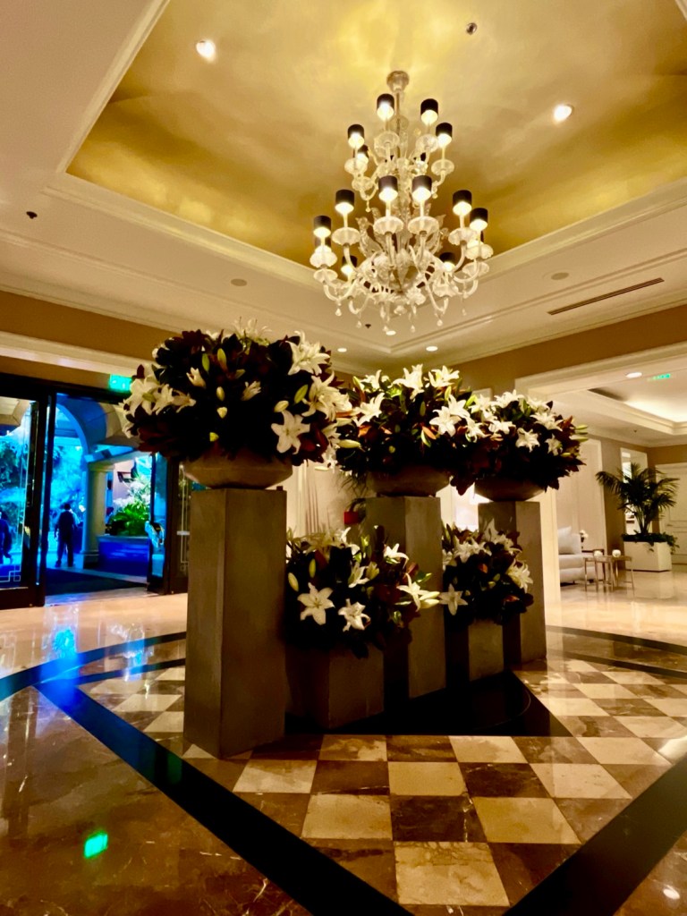 A luxurious hotel lobby featuring elegant floral arrangements, a chandelier, and polished marble flooring.