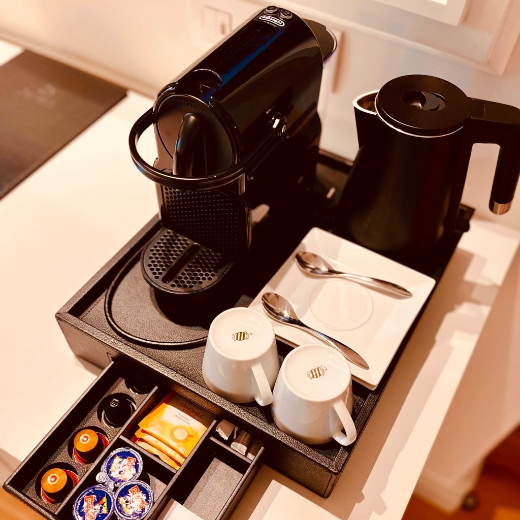 A luxury coffee and tea station featuring a black Nespresso machine, an electric kettle, two white cups, and assorted coffee pods and condiments.