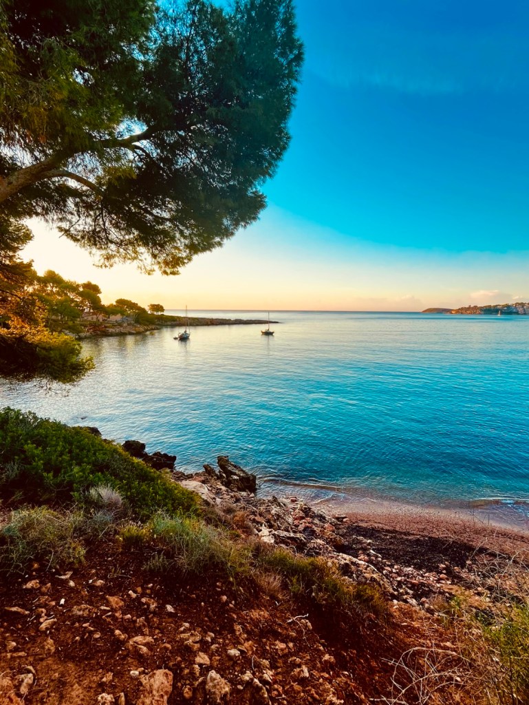 A serene view of the Mediterranean coastline at sunset, featuring a tranquil sea with boats anchored near the shore, surrounded by green trees and rocky land.