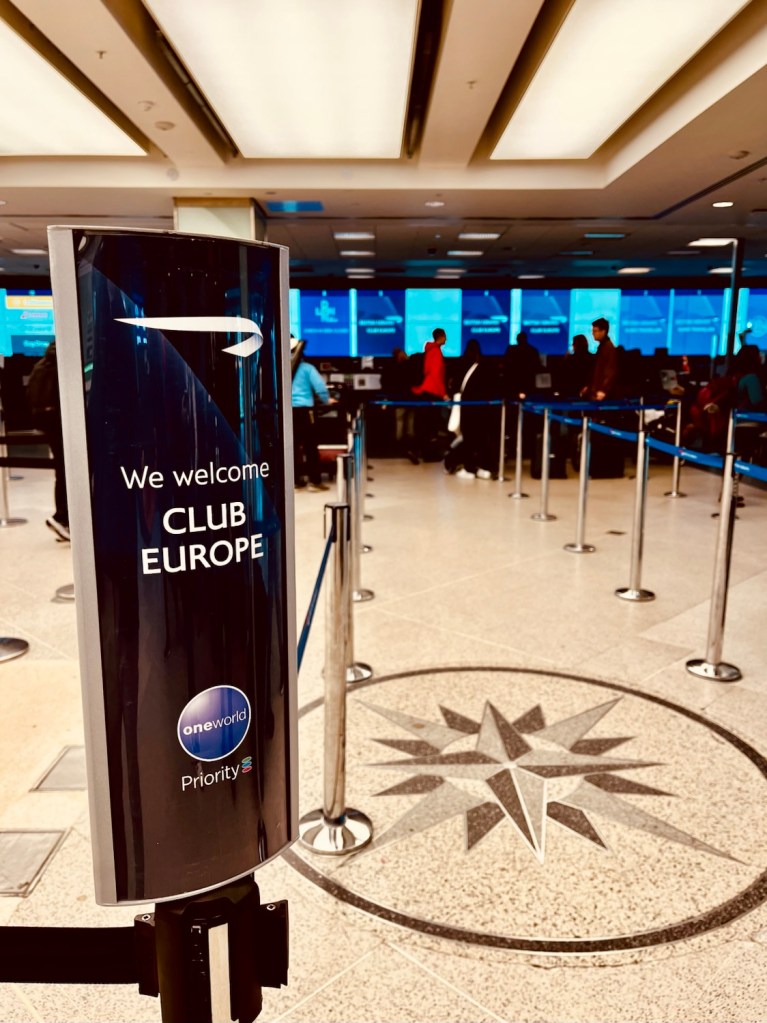 Signage welcoming passengers to British Airways Club Europe at London City Airport, with priority boarding area and check-in counters in the background.