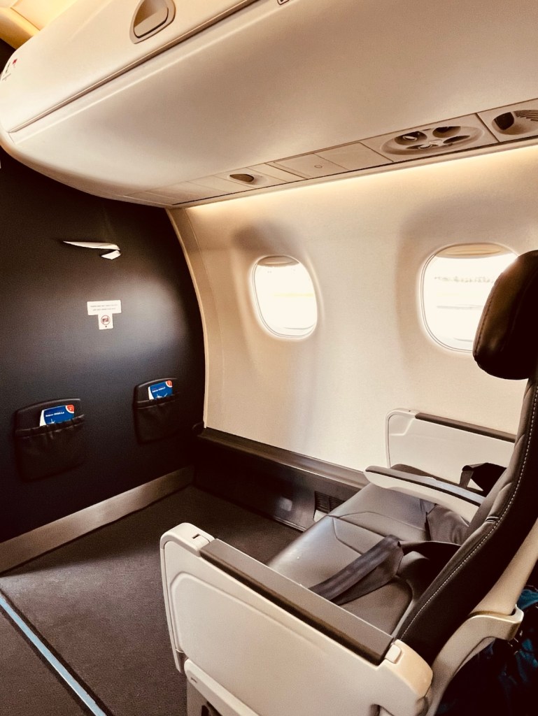 Interior view of a business class cabin on an Embraer jet, featuring a spacious seat with armrests, a window view, and overhead storage compartments.
