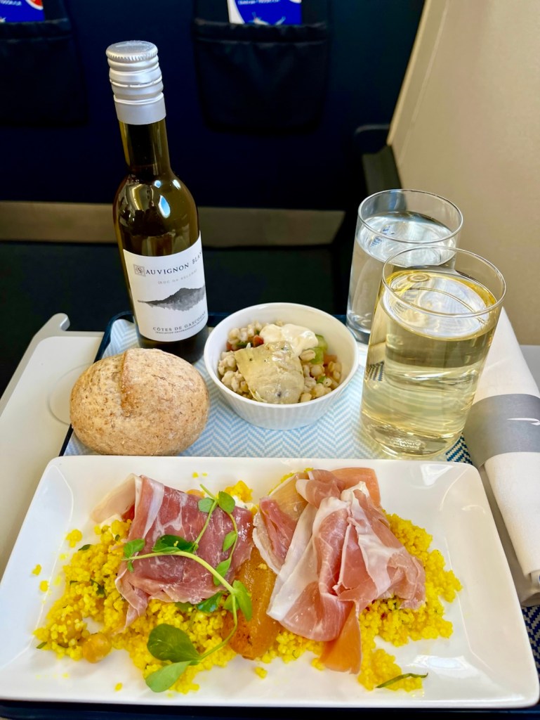 A meal tray featuring couscous, thinly sliced meat, a roll, a salad, and a bottle of white wine onboard a flight.