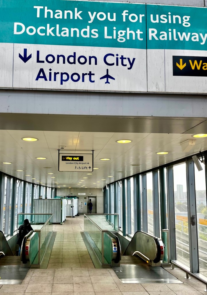 Interior view of the Docklands Light Railway station with a sign directing travelers to London City Airport, featuring escalators and glass walls.