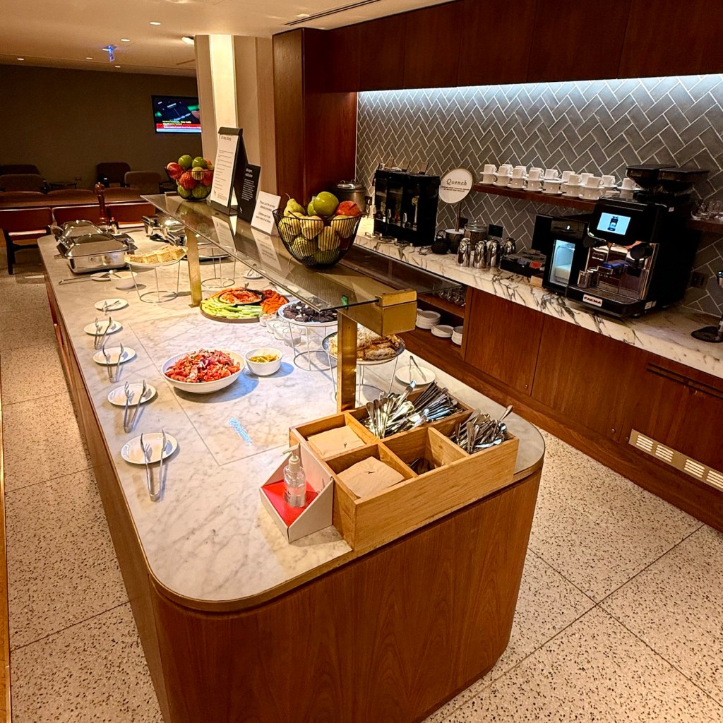 A well-organized buffet area in an airport lounge with various food items, including salads and fruits, displayed on a marble countertop, alongside a coffee station and stylish wooden accents.