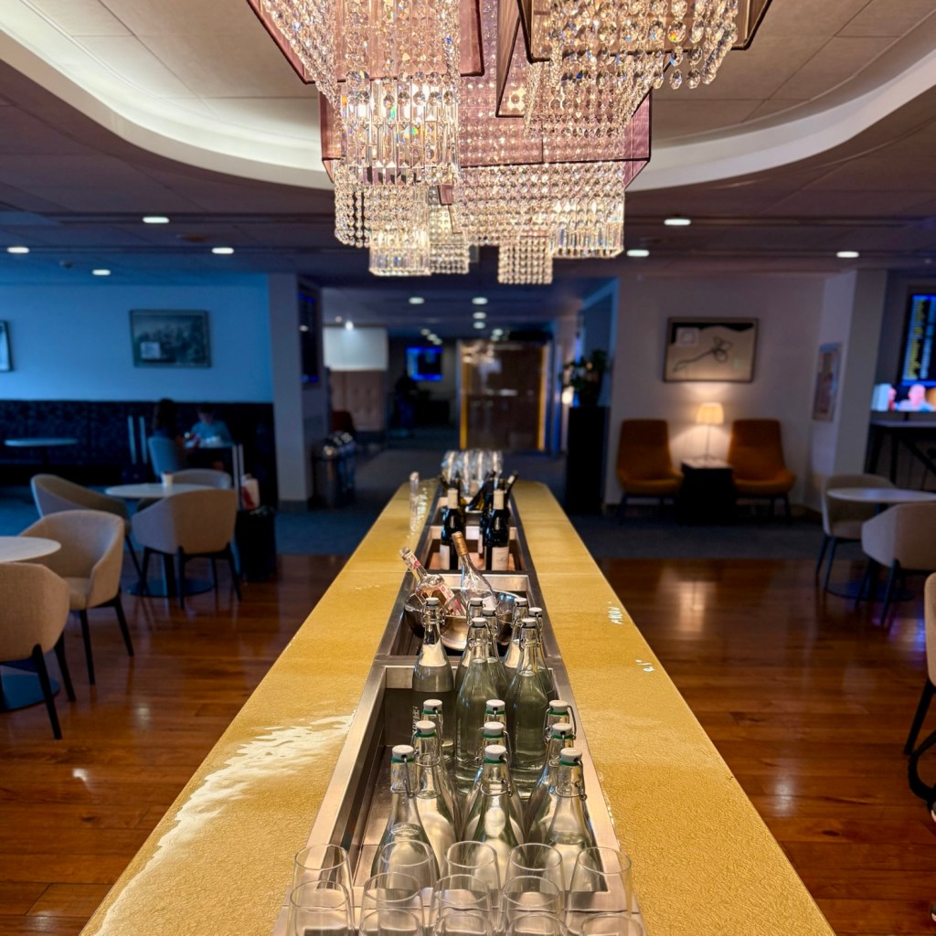 A well-lit bar area in an airport lounge featuring sparkling chandeliers and a counter with various drinks, including bottles of sparkling water and wine.