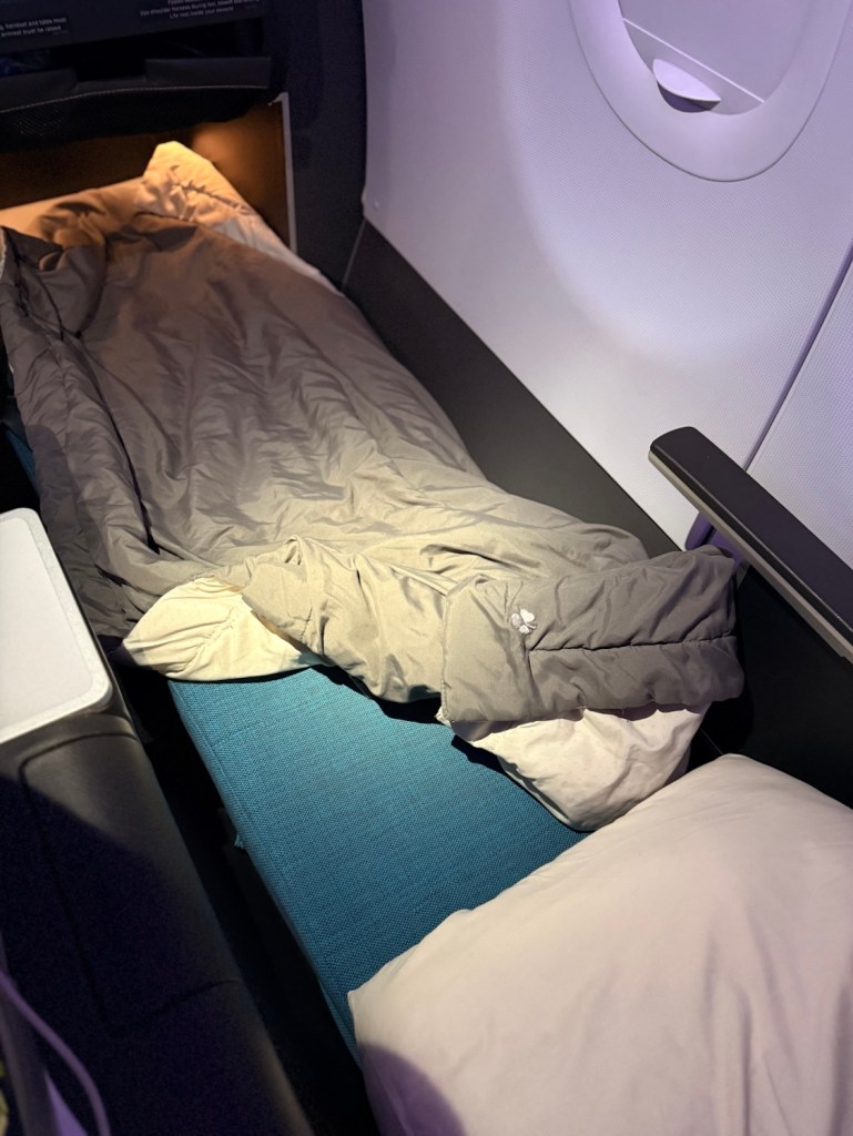 An Aer Lingus business class seat configured into a flat bed with a blanket and pillows, set against a softly lit cabin interior.