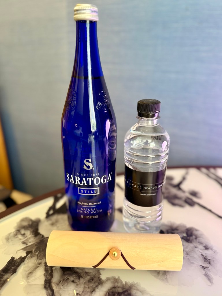 A blue Saratoga water bottle, a clear bottled water with a label indicating 'Park Hyatt Washington,' and a wooden box on a decorative table.