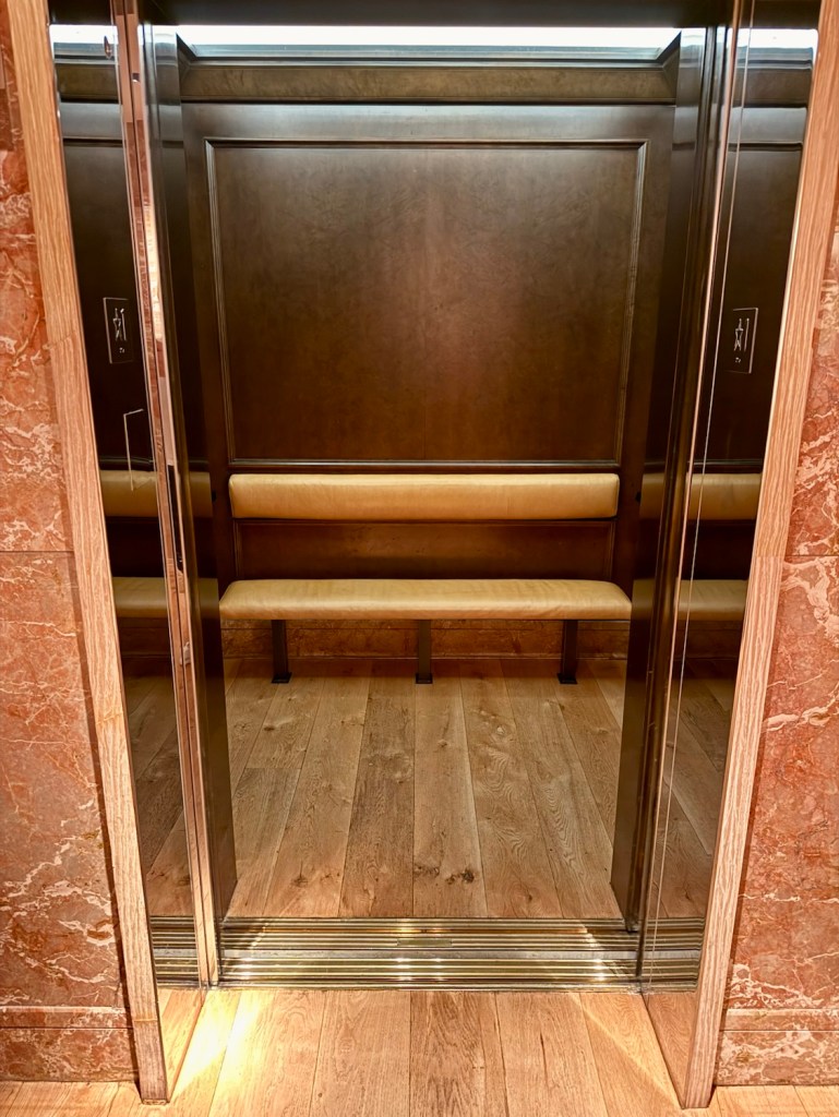 Interior of a hotel elevator featuring wooden paneling, a bench seat, and polished metal accents.