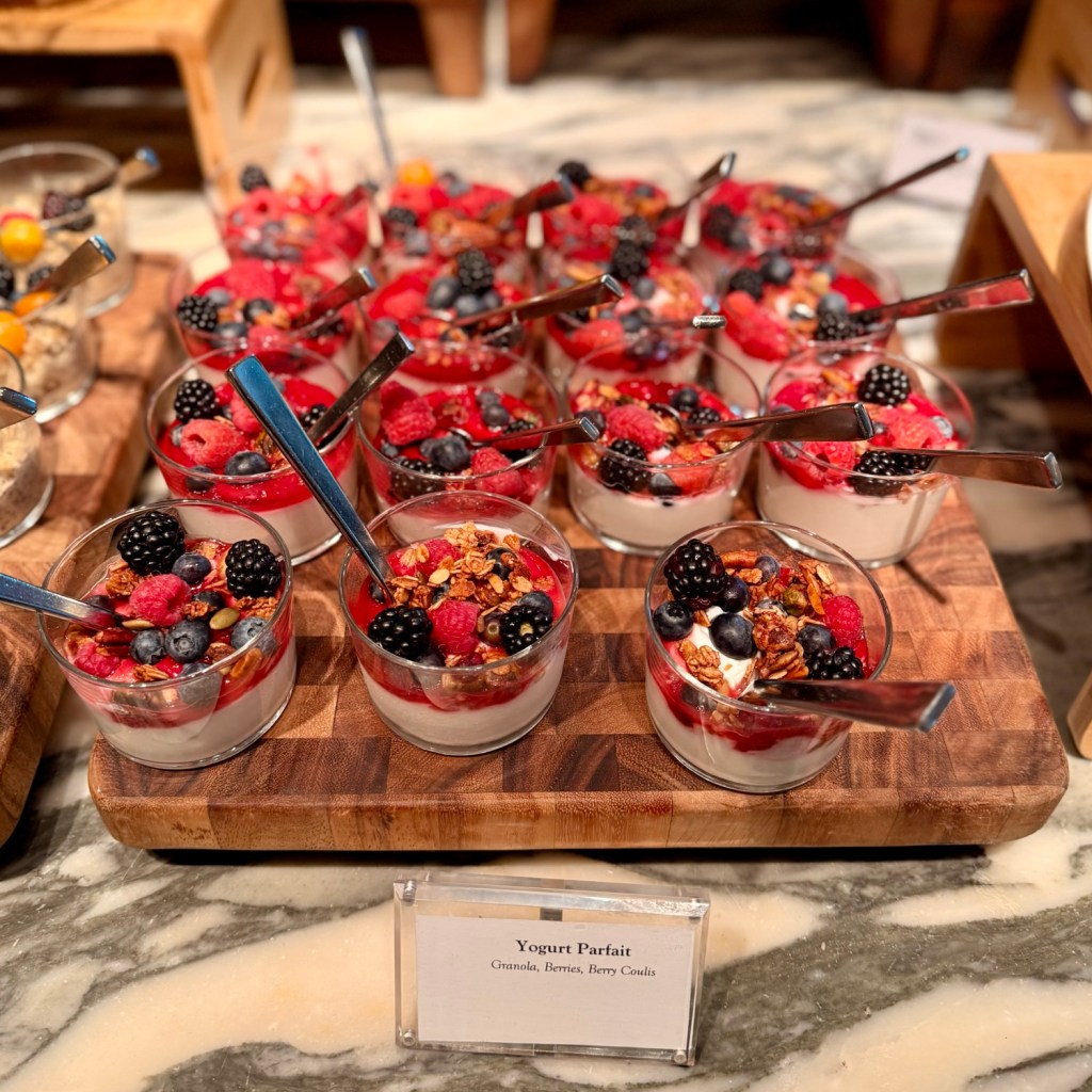 A display of yogurt parfaits topped with granola, assorted berries, and a berry coulis on a wooden platter.