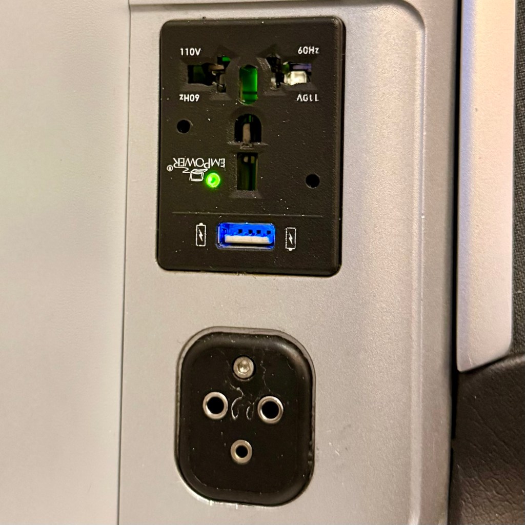 Close-up of a power outlet and USB charging port located in an airplane cabin, featuring a standard electrical socket, a USB port, and an indicator light.