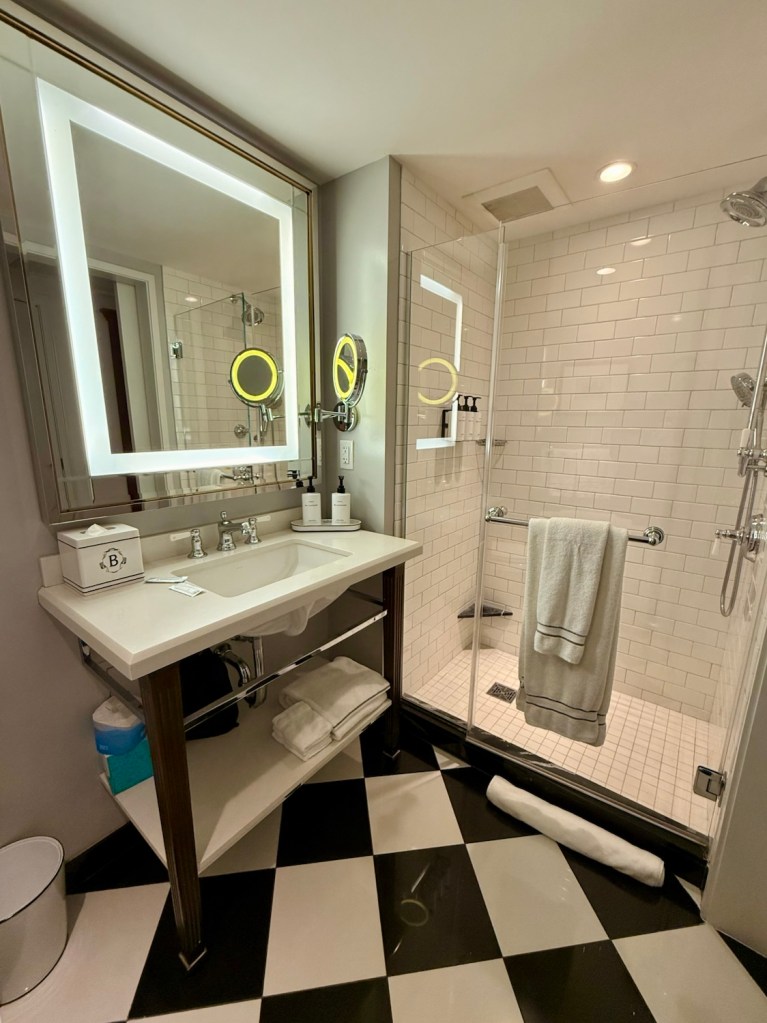 A modern hotel bathroom featuring a stylish sink with a large illuminated mirror, a glass-enclosed shower with white subway tiles, and a black and white checkered floor.