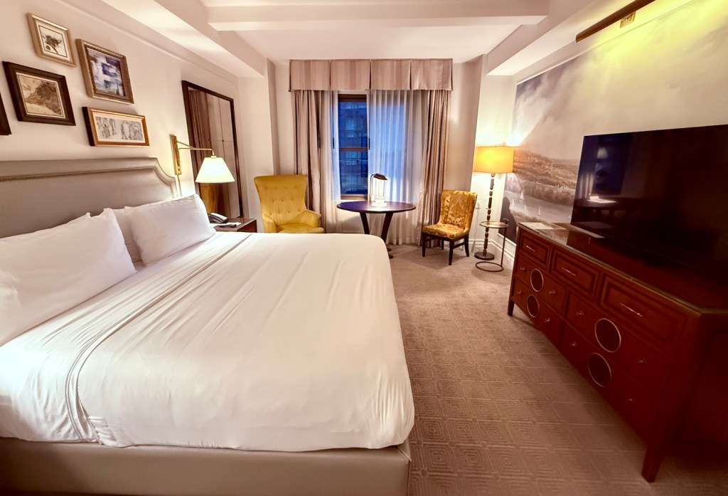 A well-appointed hotel room featuring a king-sized bed with crisp white linens, a yellow accent chair, a decorative lamp, a small table, and a large flat-screen TV, all surrounded by soft lighting and elegant decor.