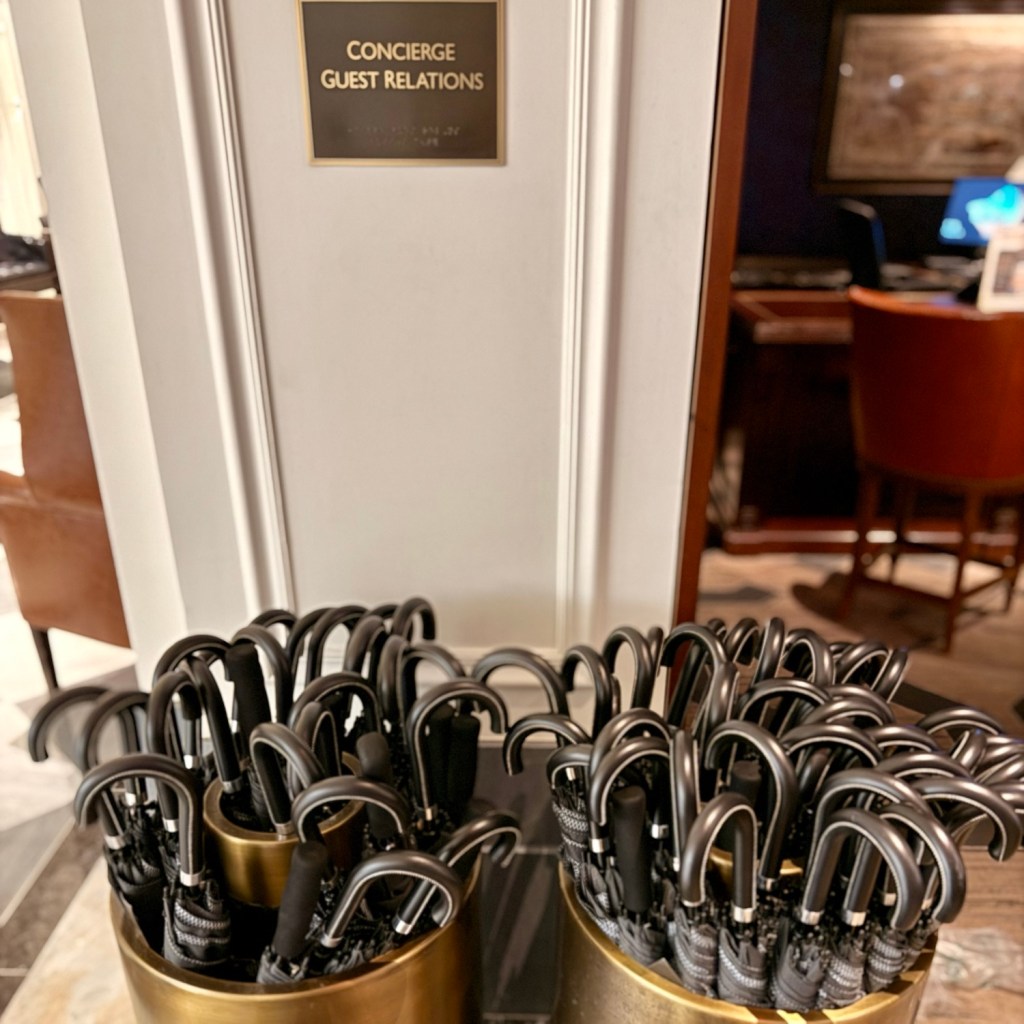 A display of black umbrellas in brass containers, located near a sign reading 'Concierge Guest Relations' in a hotel setting.