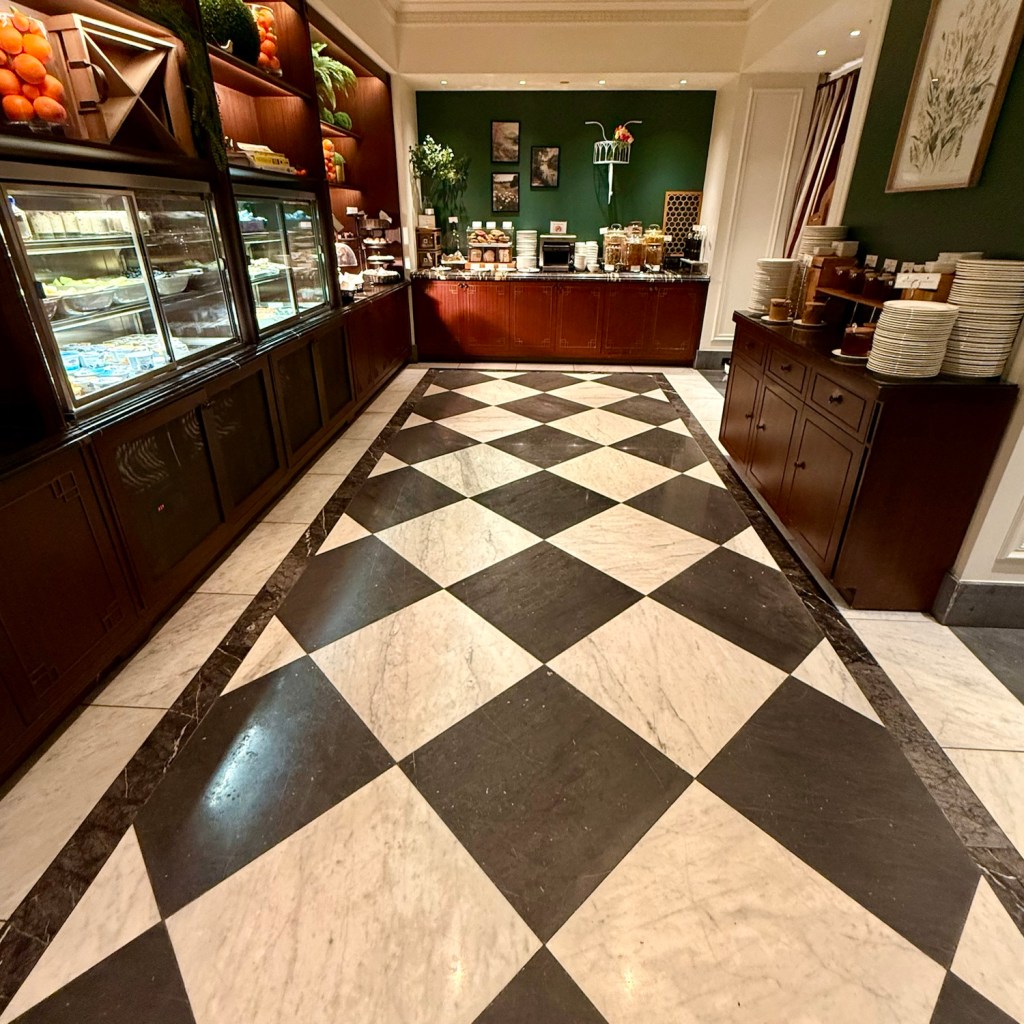 Interior view of a hotel dining area featuring a checkered marble floor and a buffet-style food service counter with various food items displayed.