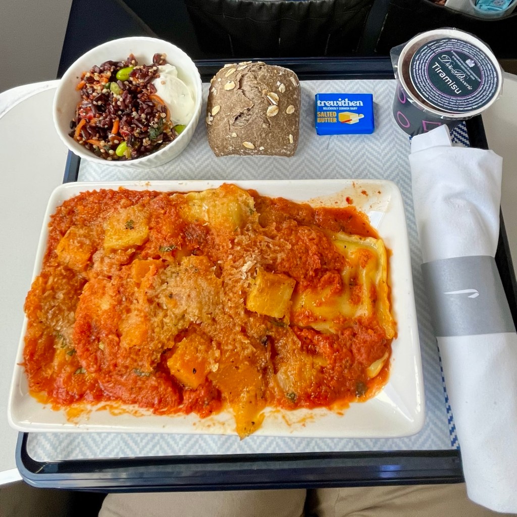 A tray of airplane food featuring a pasta dish with tomato sauce and cheese, accompanied by a bowl of salad, a piece of bread, butter, and a dessert cup.