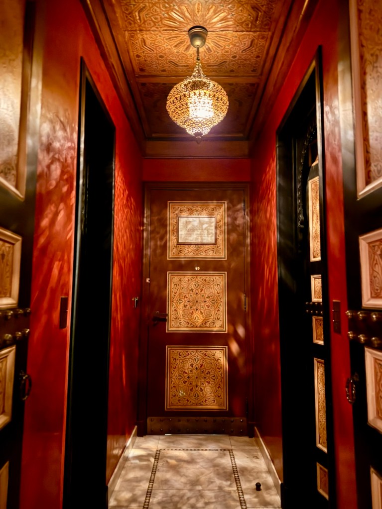 A beautifully decorated hallway showcasing ornate wooden doors, intricate ceiling designs, and a glowing hanging lantern in La Mamounia, Marrakech.