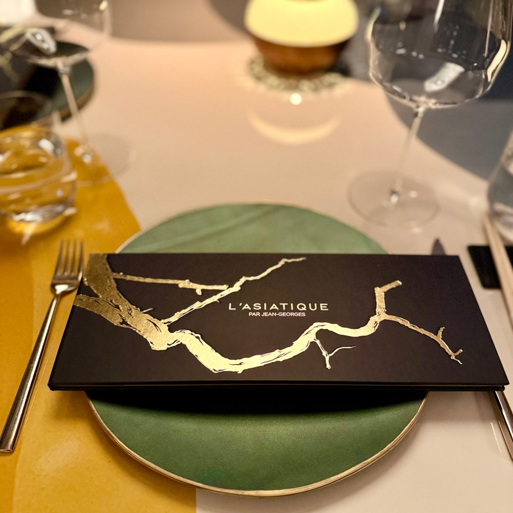 A dining table setting featuring the menu for 'L'Asiatique par Jean-Georges' on a green plate with elegant gold detailing, accompanied by a fork and wine glass.
