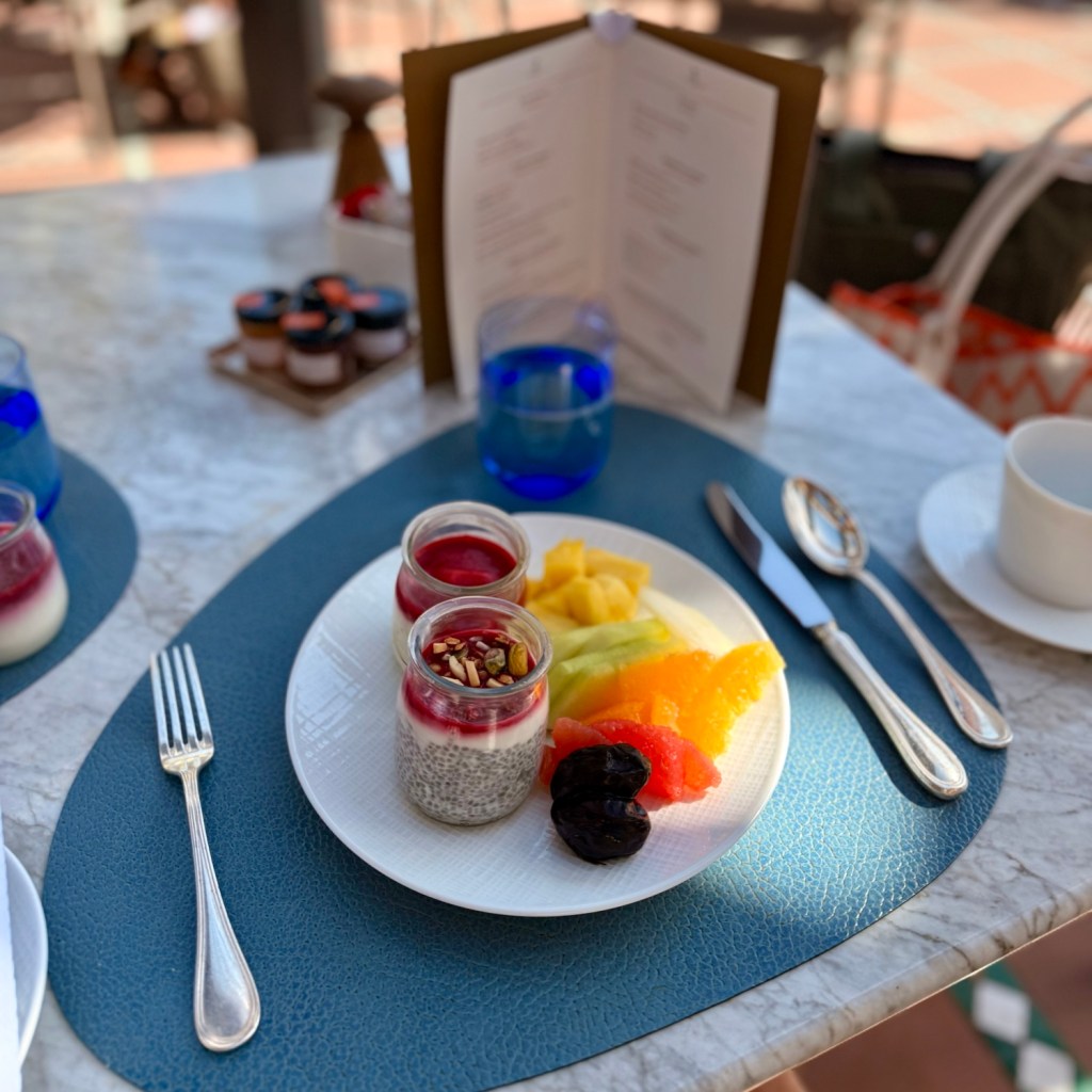 A beautifully arranged breakfast plate featuring chia pudding in jars topped with fruit compote, accompanied by a colorful selection of fresh fruits like pineapple, melon, and orange, set on a textured blue placemat beside a menu and glass of water.