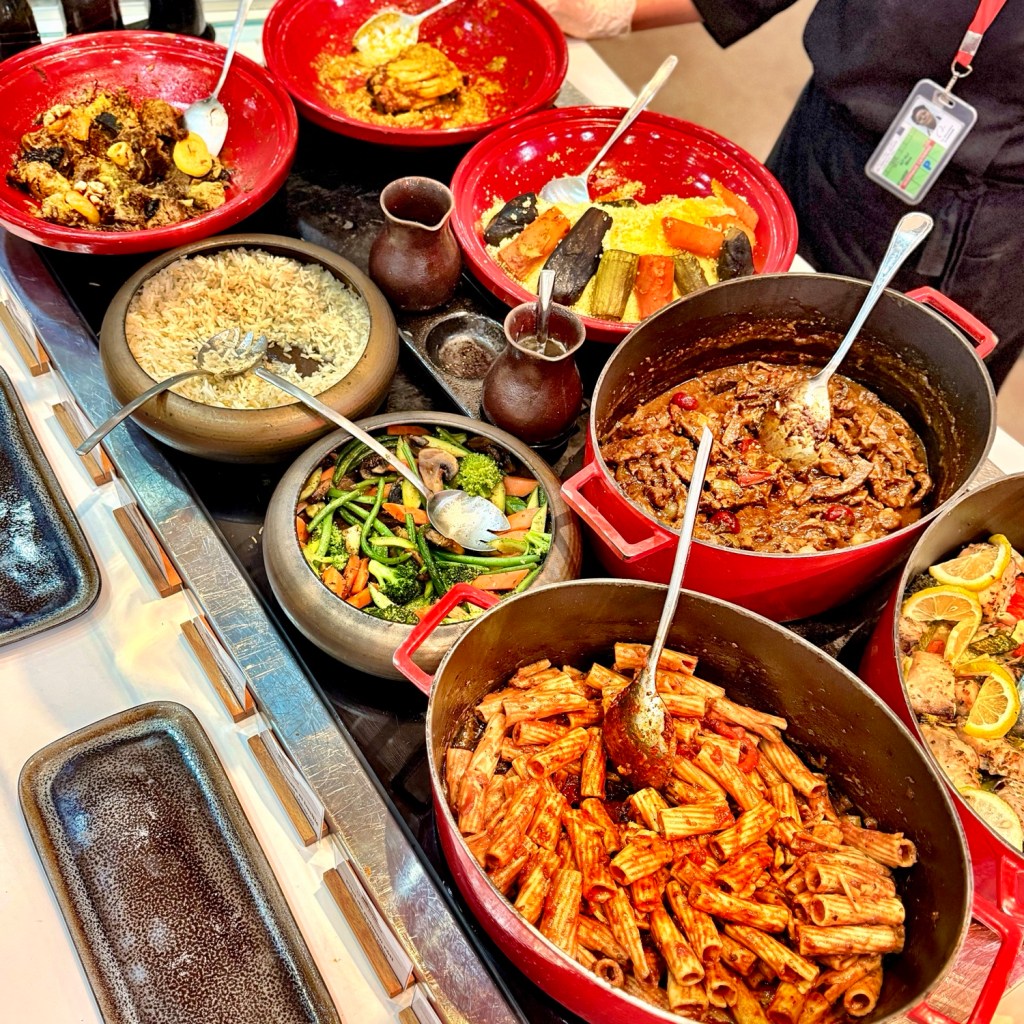 A colorful assortment of dishes on a buffet spread, including red bowls with various meats, vegetables, pasta, and rice, alongside serving spoons and traditional clay pots.