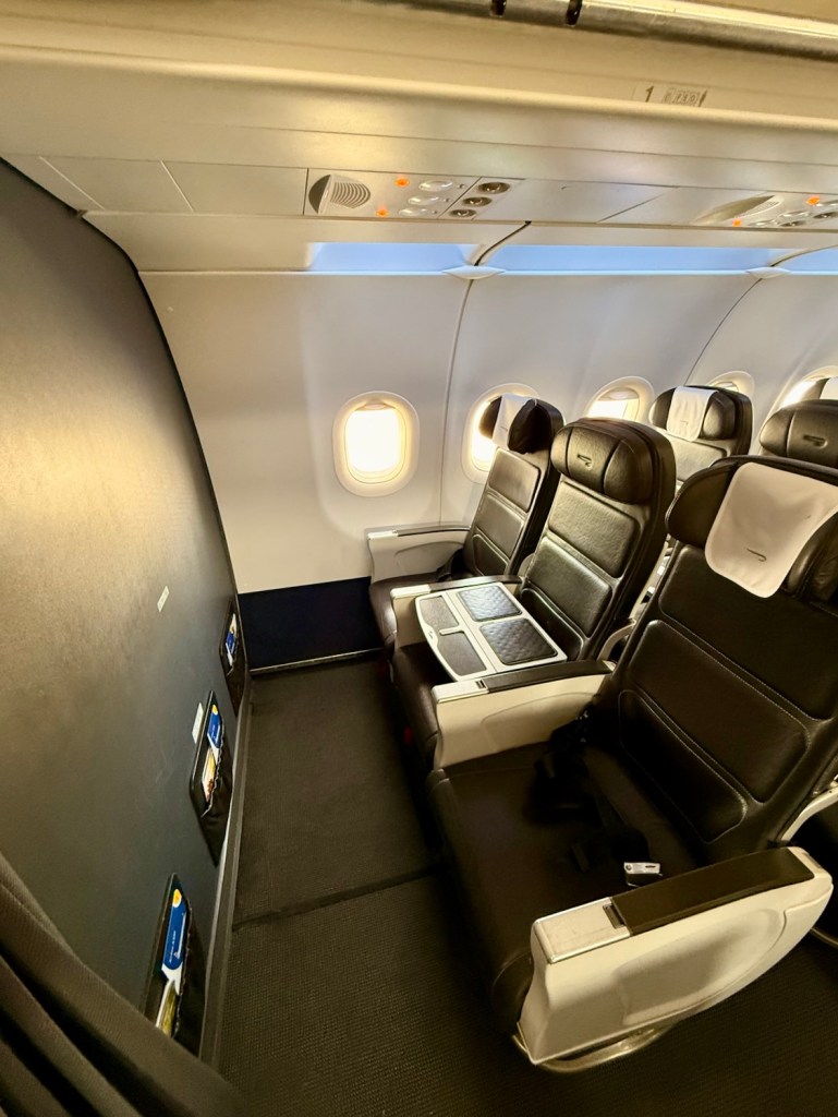 Interior view of a British Airways Airbus A320 showing the Club Europe seating arrangement, featuring two empty seats with a drinks table between them, and overhead lighting.