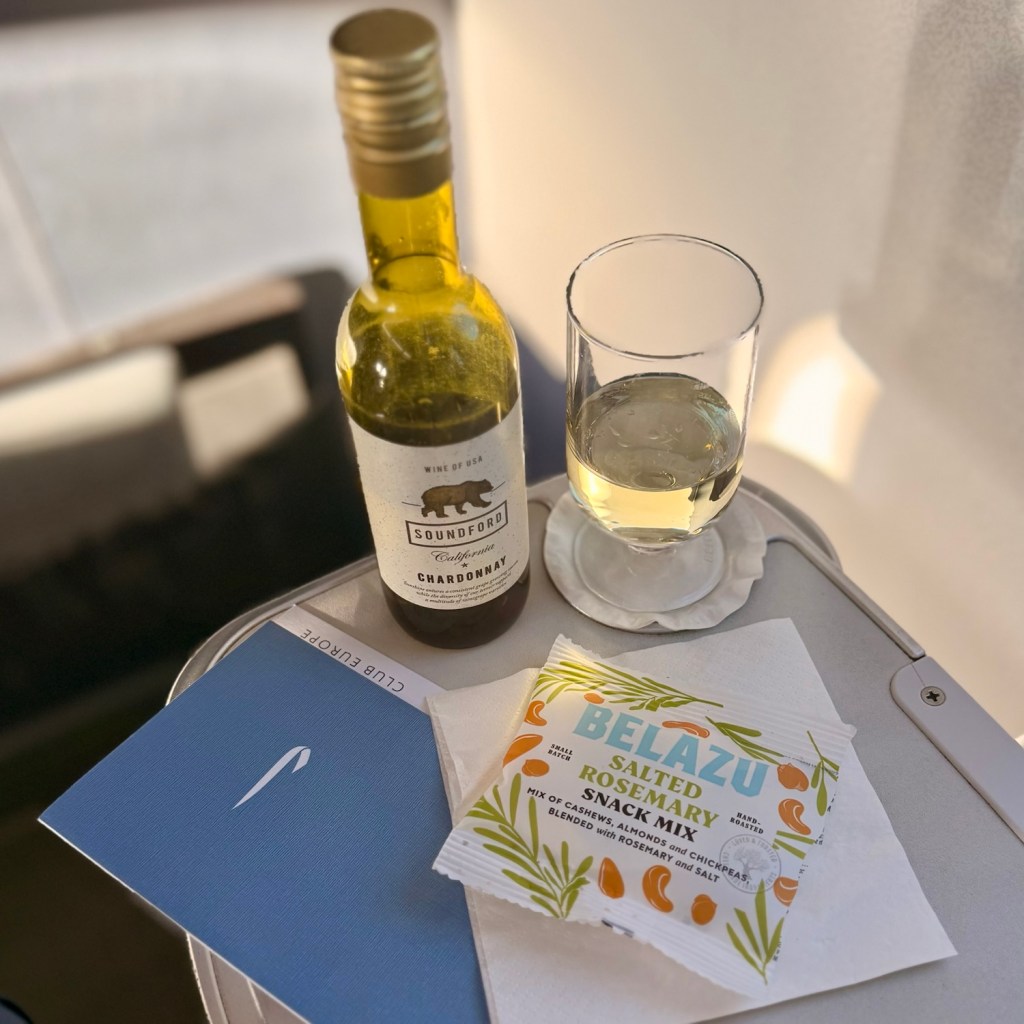 A bottle of Soundford Chardonnay wine and a glass of white wine are placed on a tray, alongside a packet of Belazu salted rosemary snack mix and a menu titled 'Club Europe'.