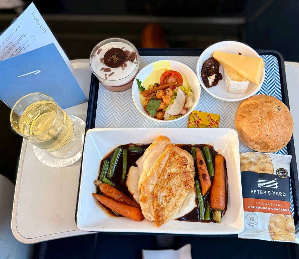 A serving tray featuring British Airways' Club Europe dinner, including roast chicken with vegetables in gravy, a side salad, a dessert cup, cheese plate, a bread roll, and a glass of white wine.