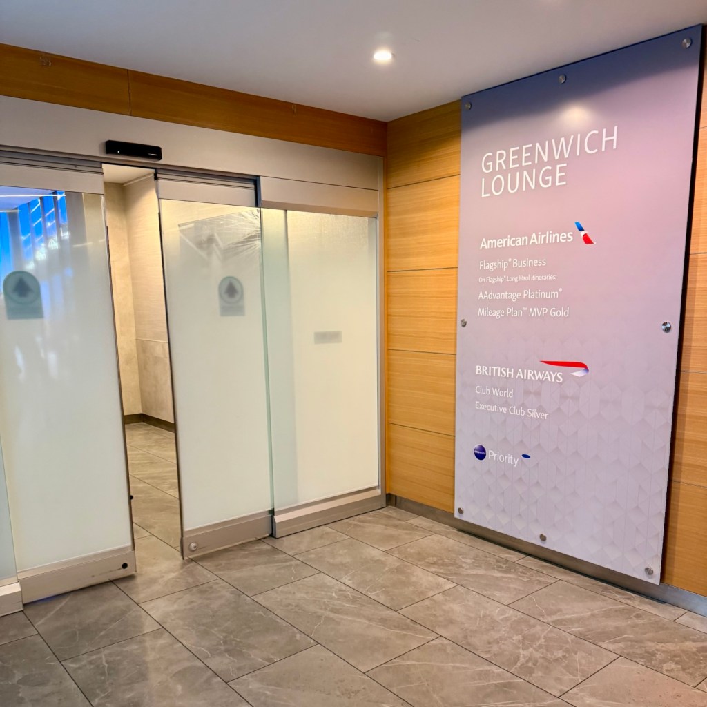 Entrance to the Greenwich Lounge at JFK Airport, featuring double doors and informational signage for American Airlines and British Airways members.