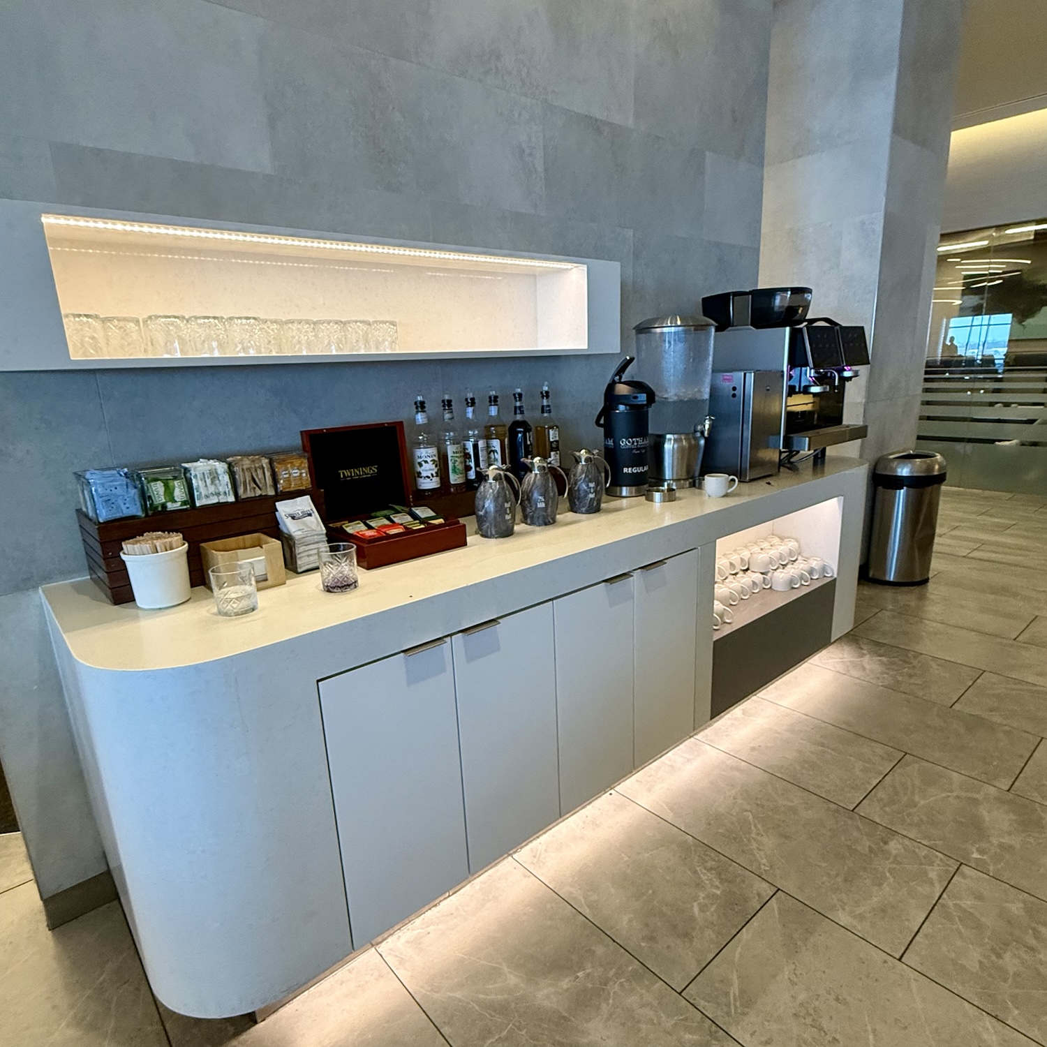 A sleek coffee and beverage station located in an airport lounge, featuring a variety of teas, coffee machines, and glassware on display, with a clean and modern aesthetic.