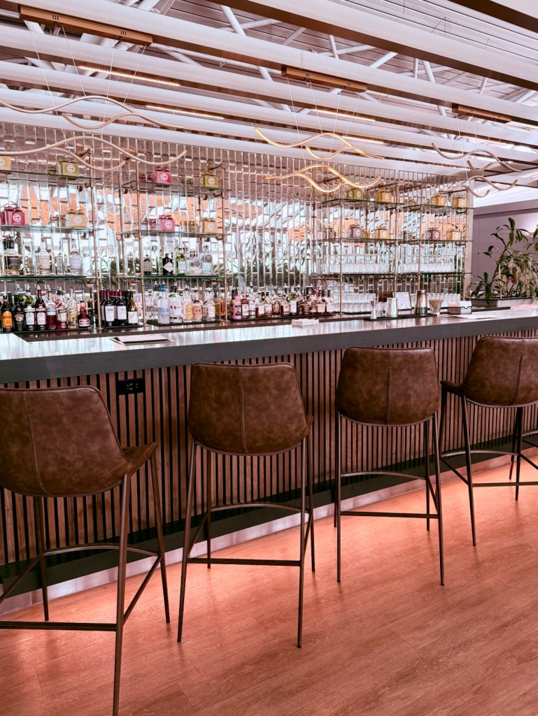 A sleek, modern bar area featuring high stools, stylish glassware, and a well-stocked liquor shelf illuminated by soft lighting.