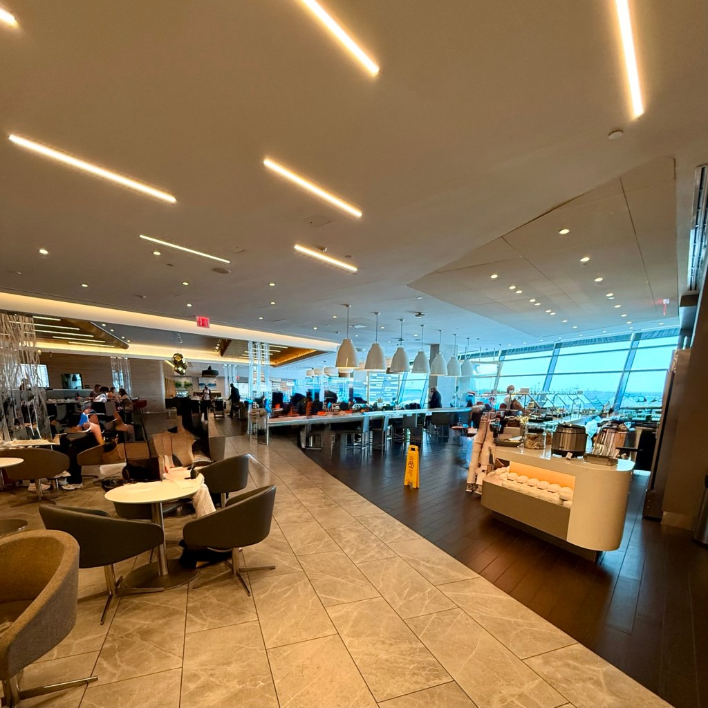 Interior view of a spacious airport lounge with modern design, featuring large windows, stylish seating areas, and a buffet service area.