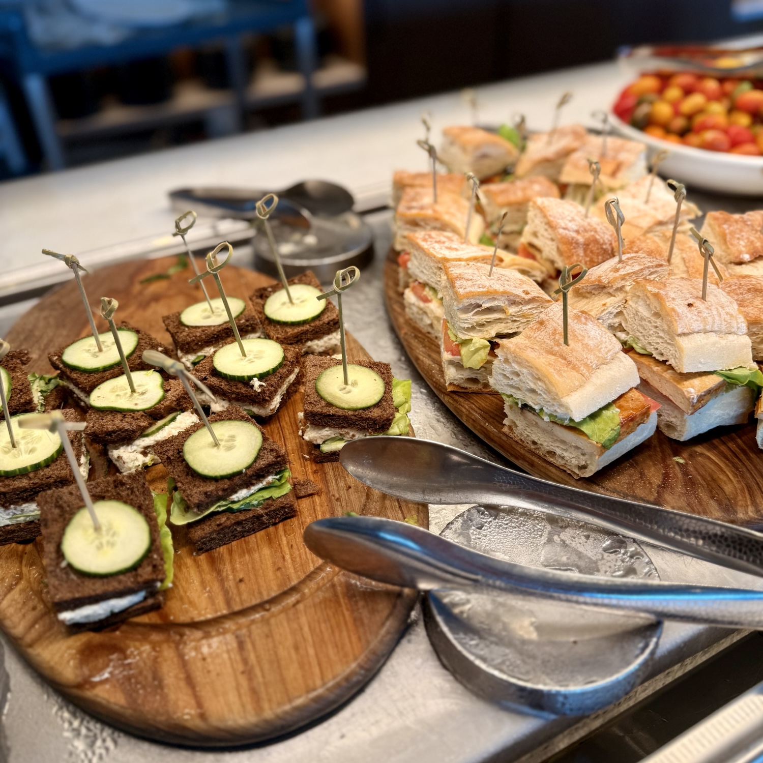 A close-up of a wooden platter featuring assorted sandwiches, including square sandwiches with cucumber slices and larger cut sandwiches, all elegantly presented with decorative picks.