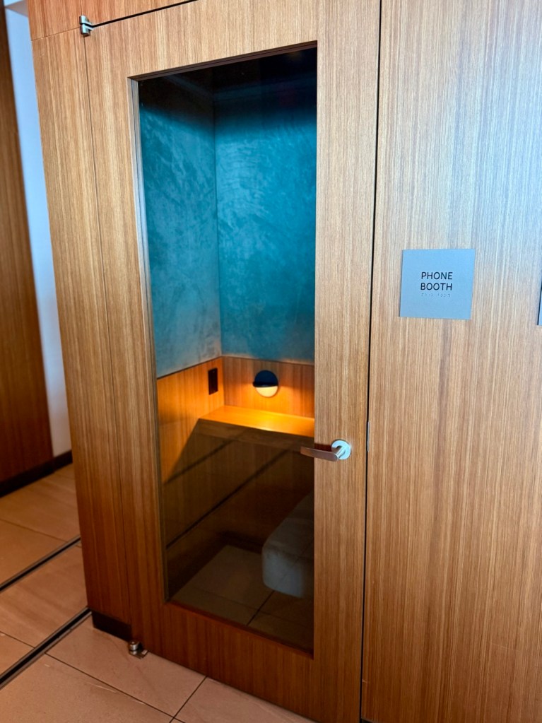 A phone booth in a lounge, featuring wooden paneling, soft lighting, and a glass door.
