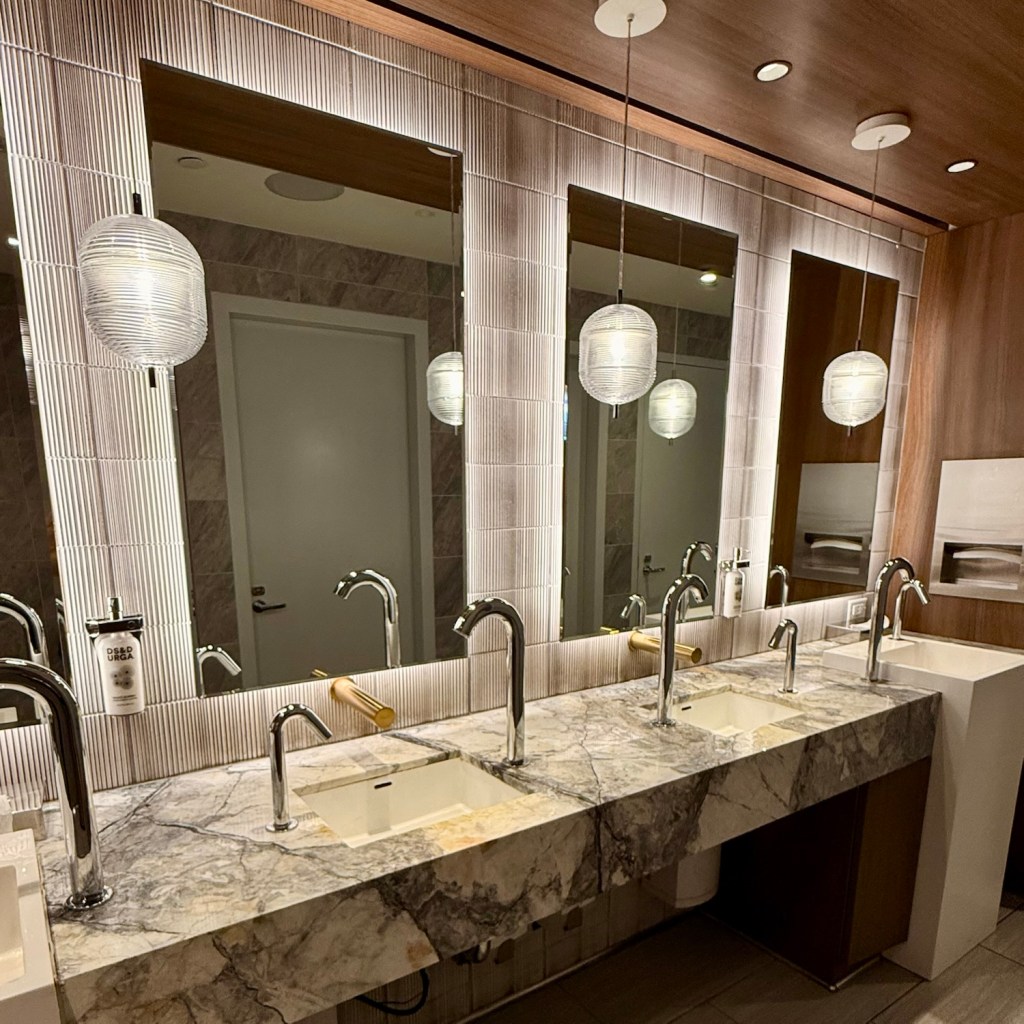 A modern airport lounge bathroom featuring sleek marble countertops, multiple sinks, stylish lighting fixtures, and large mirrors, providing a clean and elegant ambiance.