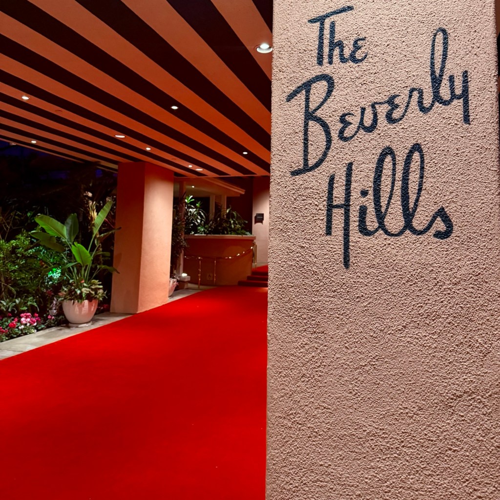Entrance to The Beverly Hills Hotel showcasing a red carpet and tropical plants in an elegant, softly lit setting.