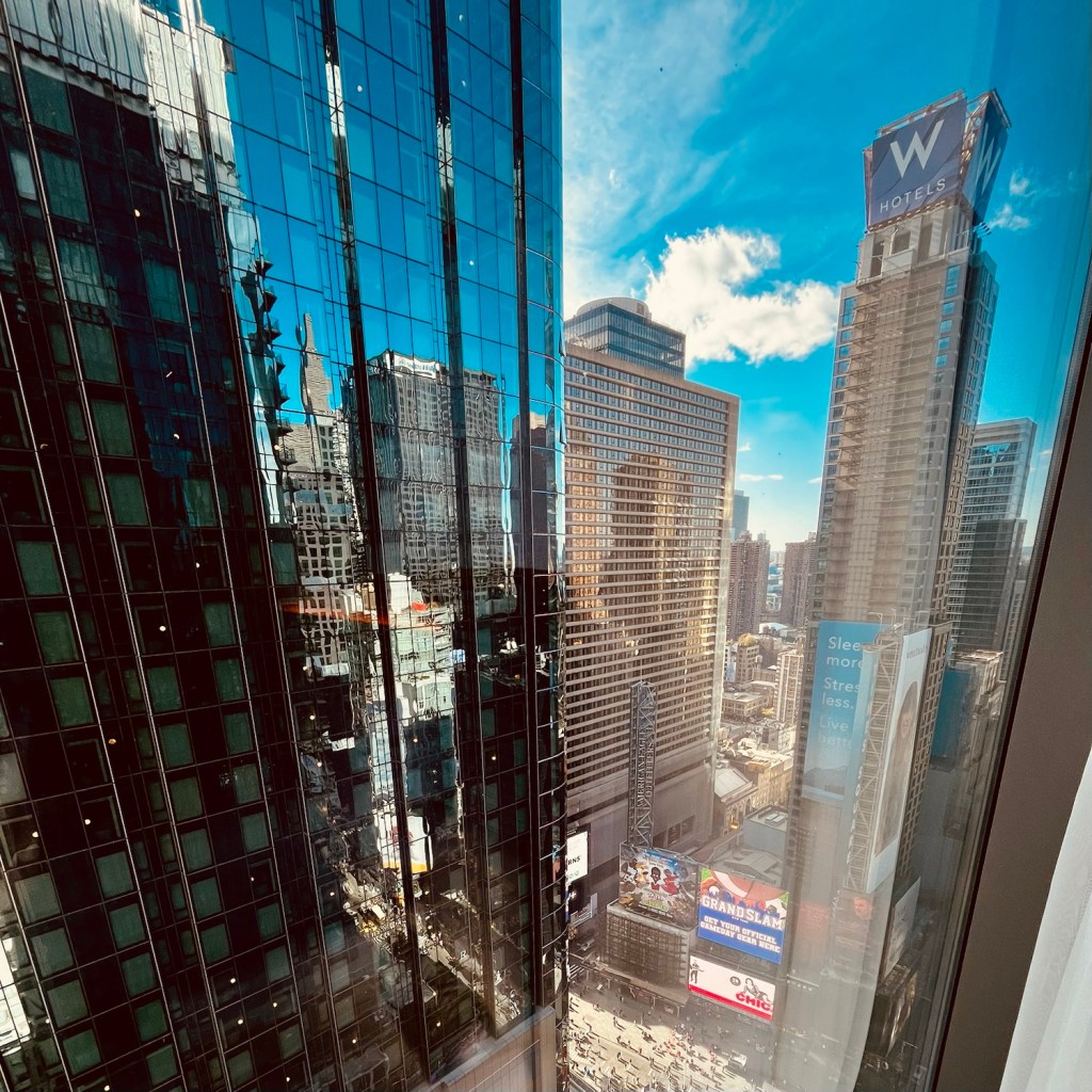 A view from a hotel window showcasing the bustling Times Square below, with tall glass buildings reflecting the blue sky and clouds.