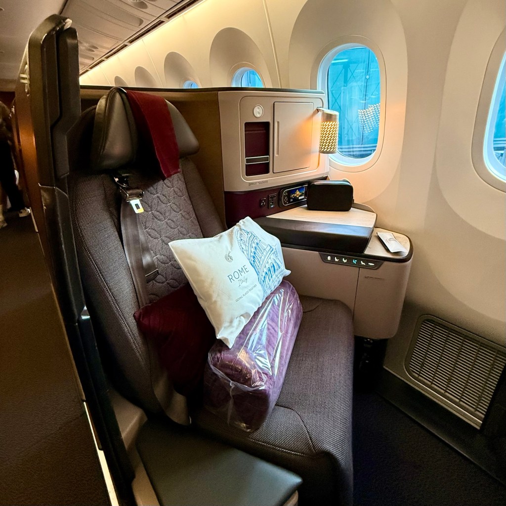 Interior view of a Qatar Airways Boeing 787-9 Business Class suite, featuring a plush seat with a decorative pillow, a small table, and large windows.