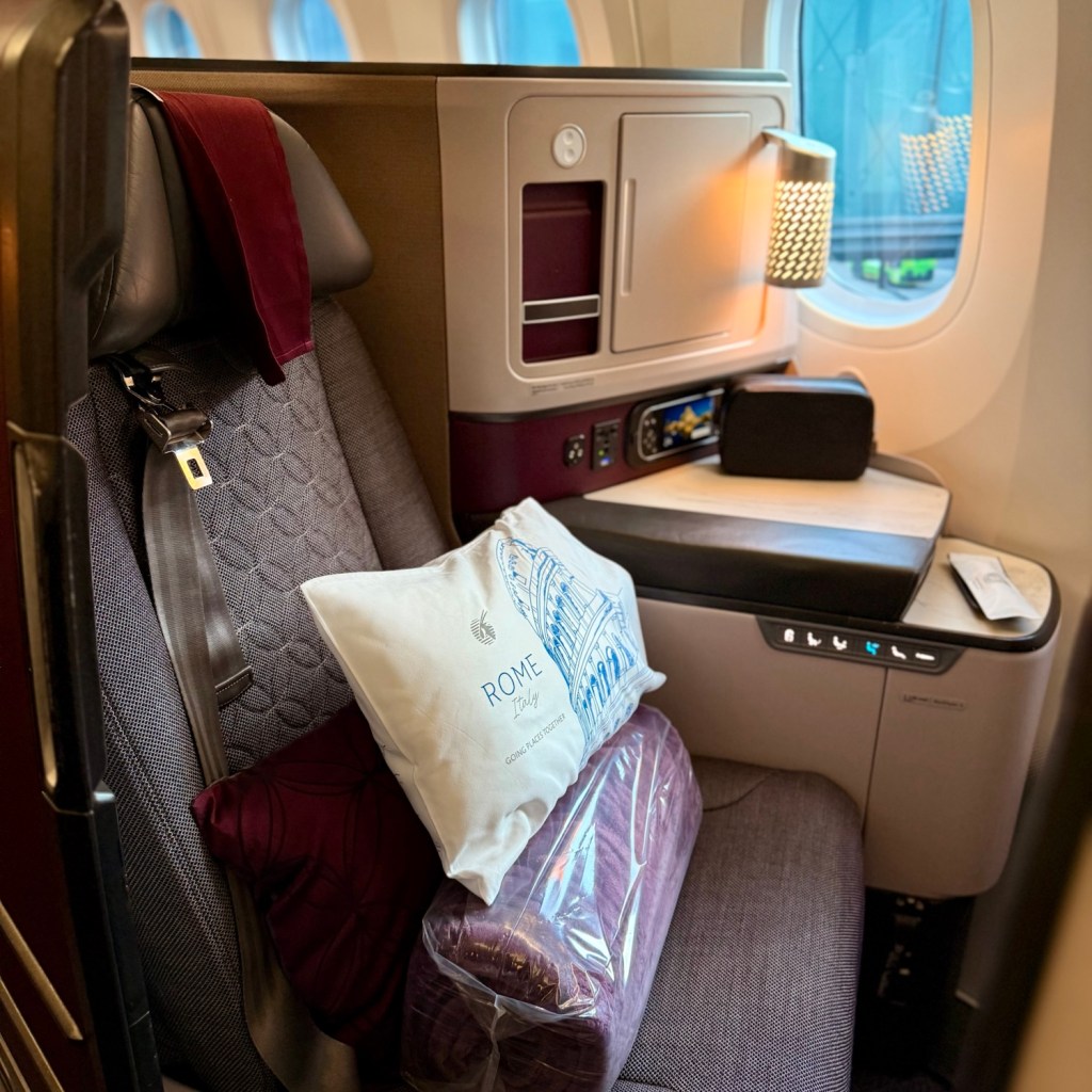 Interior view of a Qatar Airways Business Class seat on a Boeing 787-9, featuring plush seating, a decorative pillow with 'Rome' printed on it, and a cozy blanket.