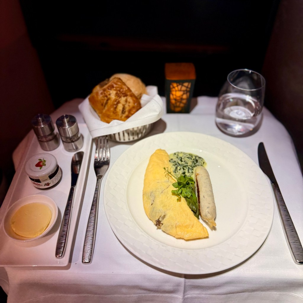 An elegantly set dining table featuring a gourmet omelette, sausage, and spinach, accompanied by bread rolls and condiments, all presented on a white tablecloth with a decorative candle in the background.