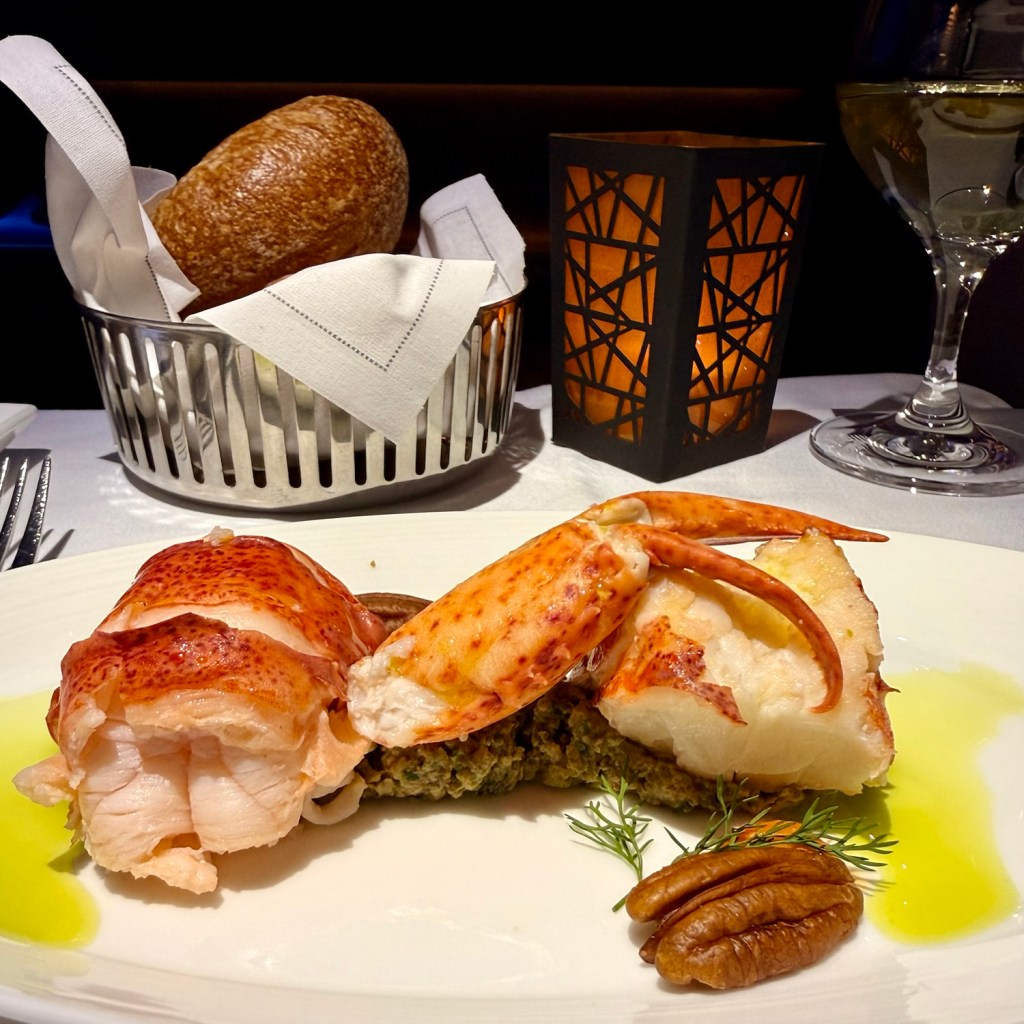 A gourmet dish featuring lobster and crab served on a white plate, accompanied by a serving of bread in a silver basket, a lit candle, and a glass of white wine.