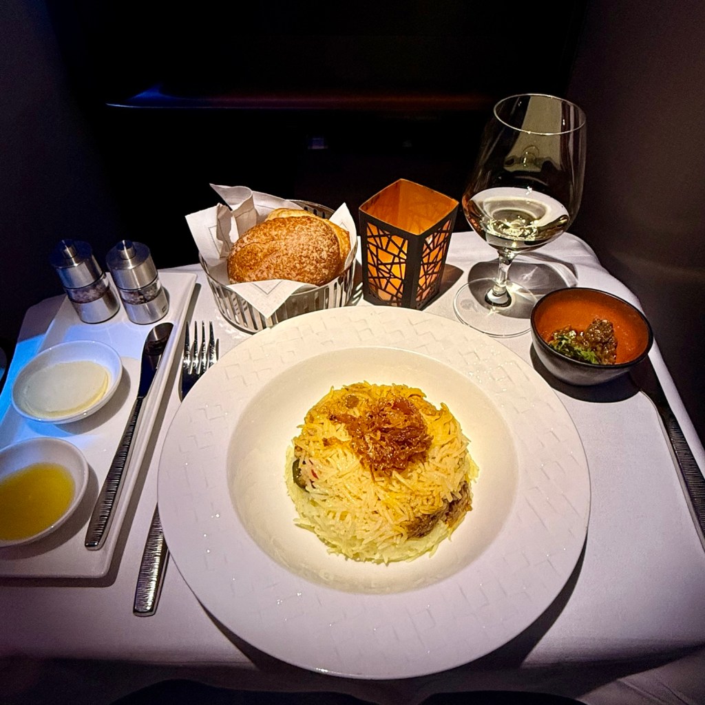 A beautifully arranged plate of rice topped with crispy onions, accompanied by a side of sauce, a roll of bread, and a glass of white wine, all set on a table with elegant tableware.
