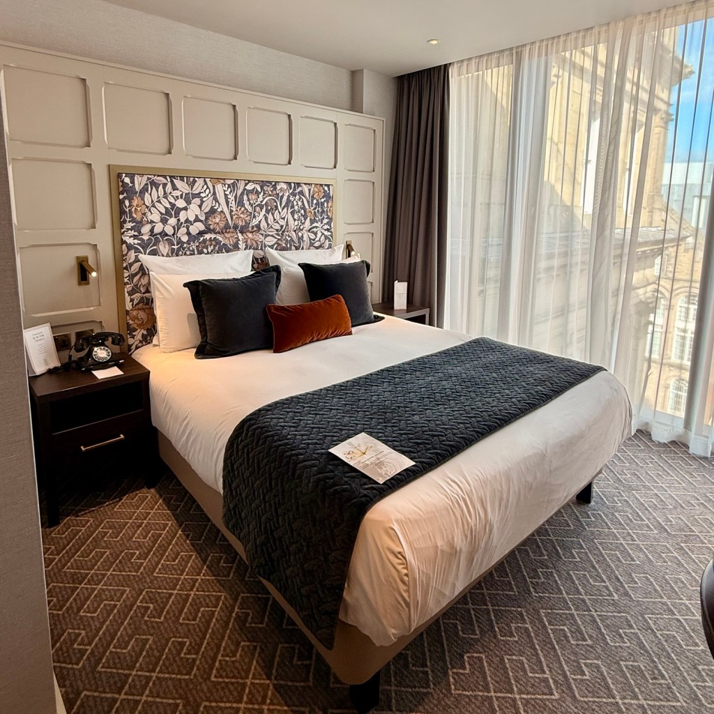 A modern hotel room featuring a queen-sized bed with decorative pillows, a dark blanket, a bedside table with a phone, and large windows with sheer curtains allowing natural light.