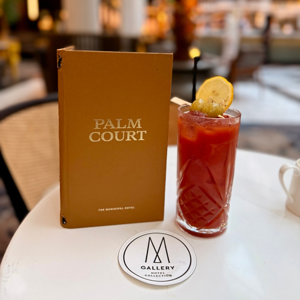 A close-up of a cocktail glass filled with a red drink, accompanied by a lemon slice and garnish, with a menu labeled 'Palm Court' from the Municipal Hotel in the background and a branded coaster.