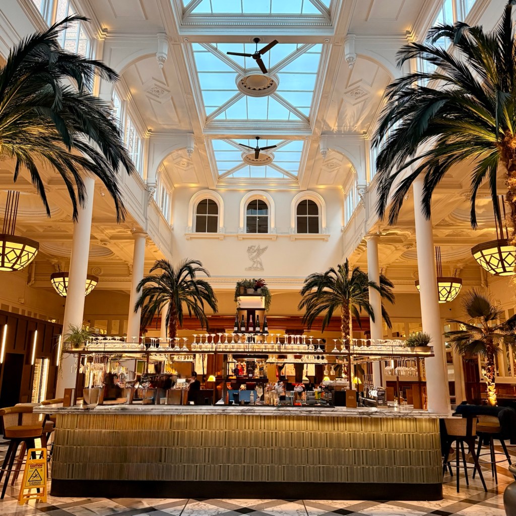 Interior view of the Palm Court Atrium featuring tall palm trees, a stylish bar with illuminated shelves, and large skylights, showcasing a blend of historic and contemporary design.