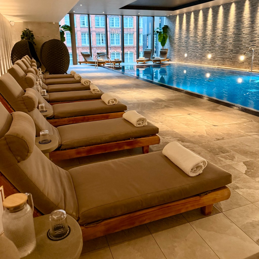 Interior view of a spa area featuring lounge chairs and an indoor swimming pool, with large windows allowing natural light to flood the space.