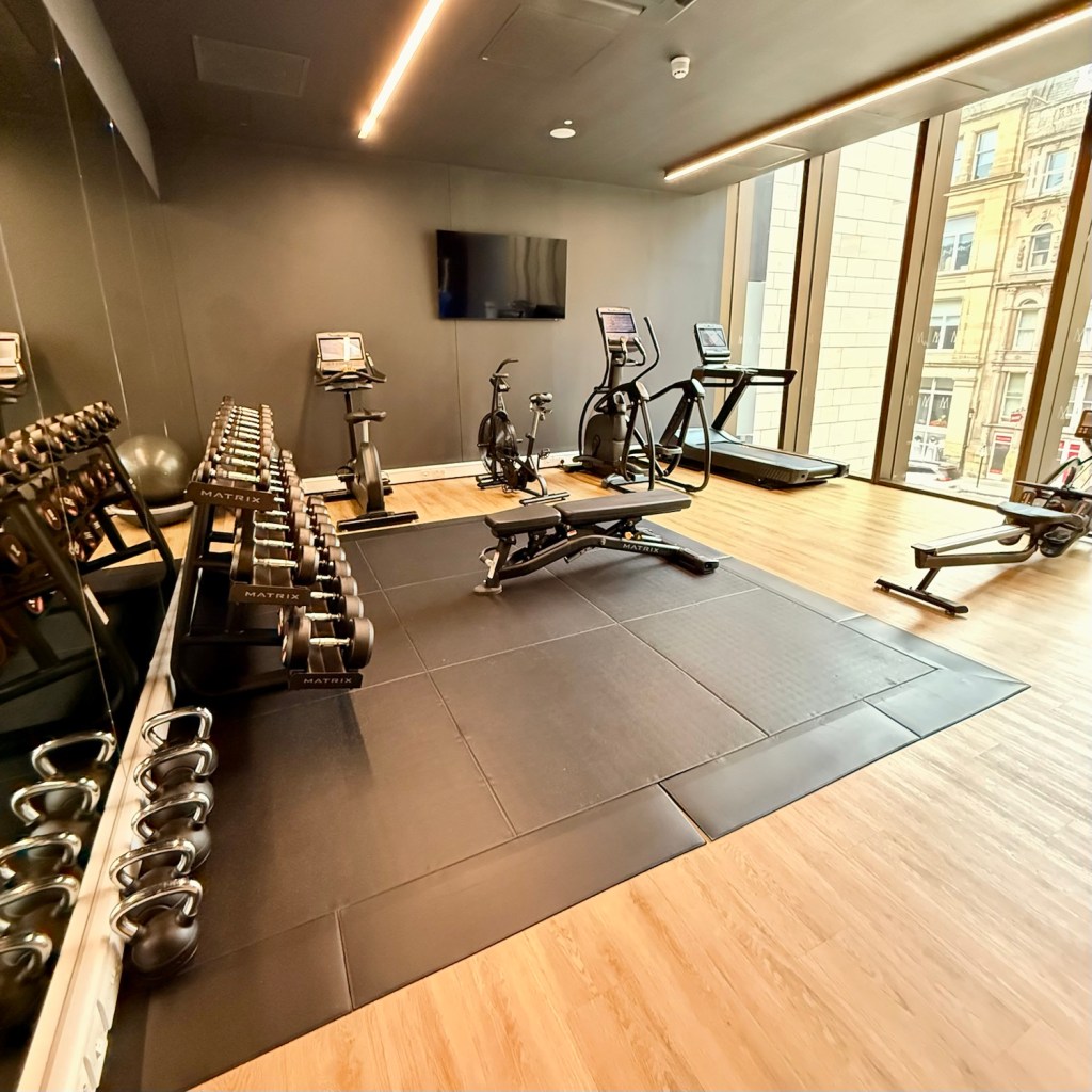 A modern gym with exercise equipment, including treadmills, elliptical machines, dumbbells, and a workout bench, set against large windows providing natural light.